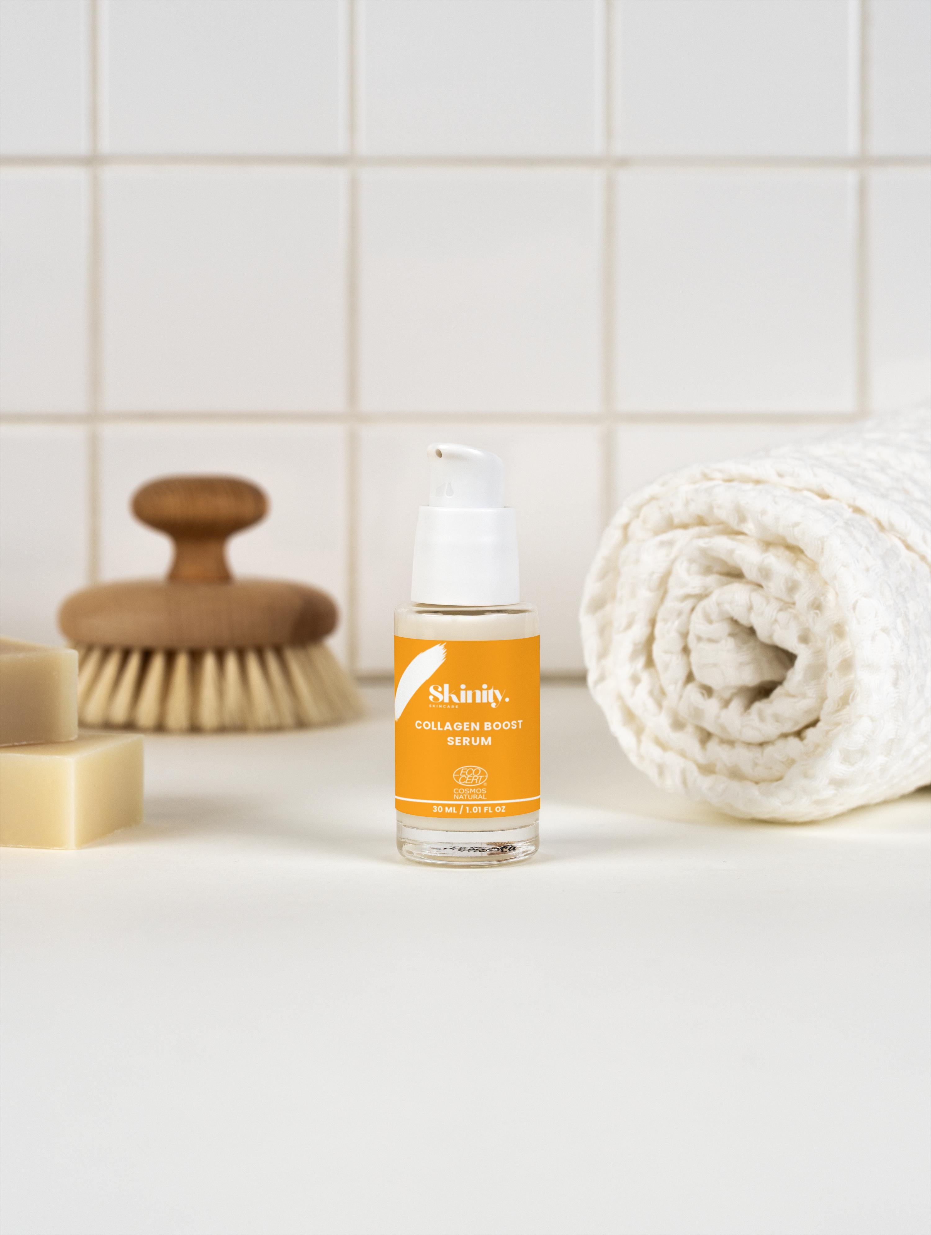 Collagen Boost Serum for All Skin positioned with a towel, soap, and a brush on a white background.
