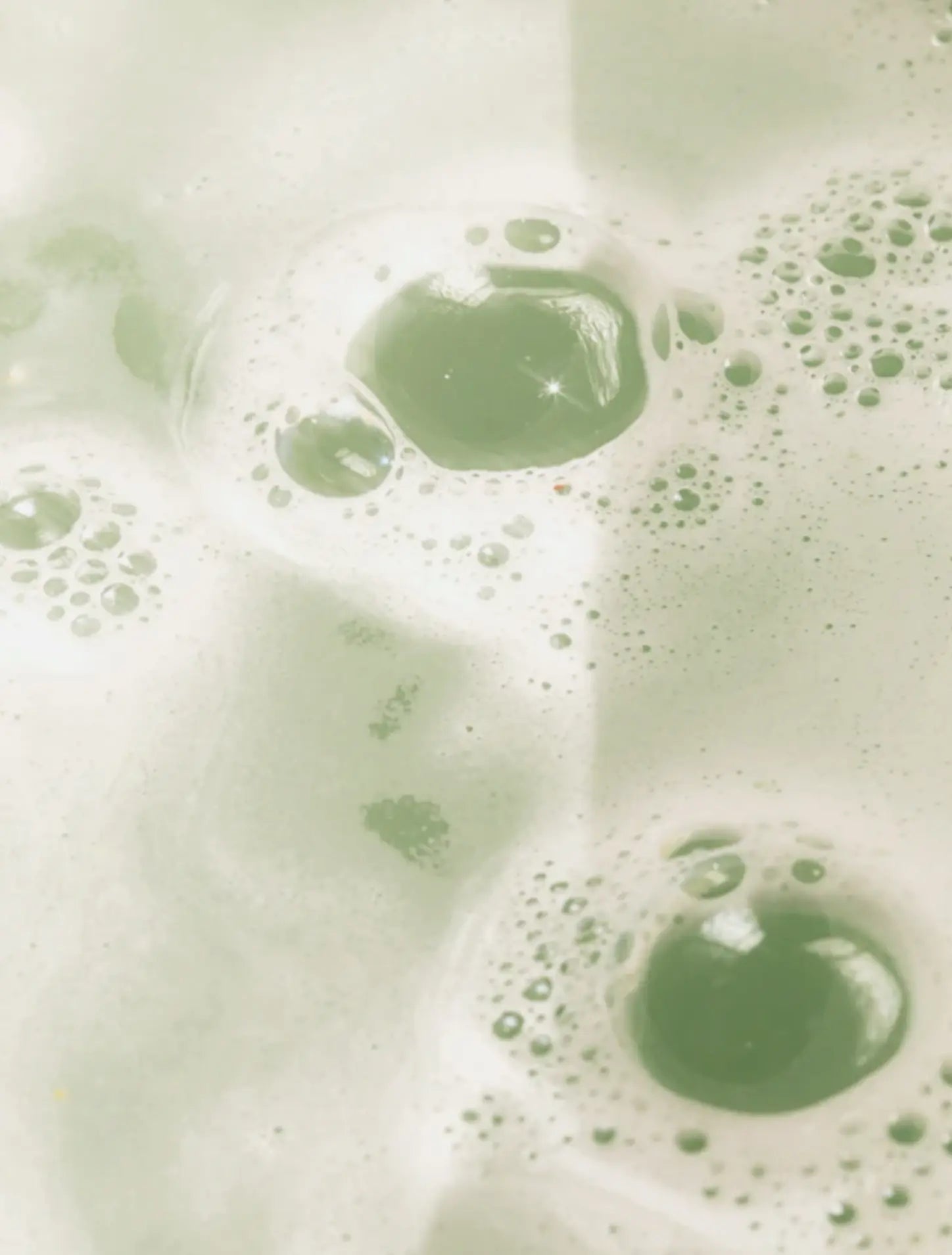A close-up abstract view of pale mint green liquid with frothy bubbles and translucent oval highlights.