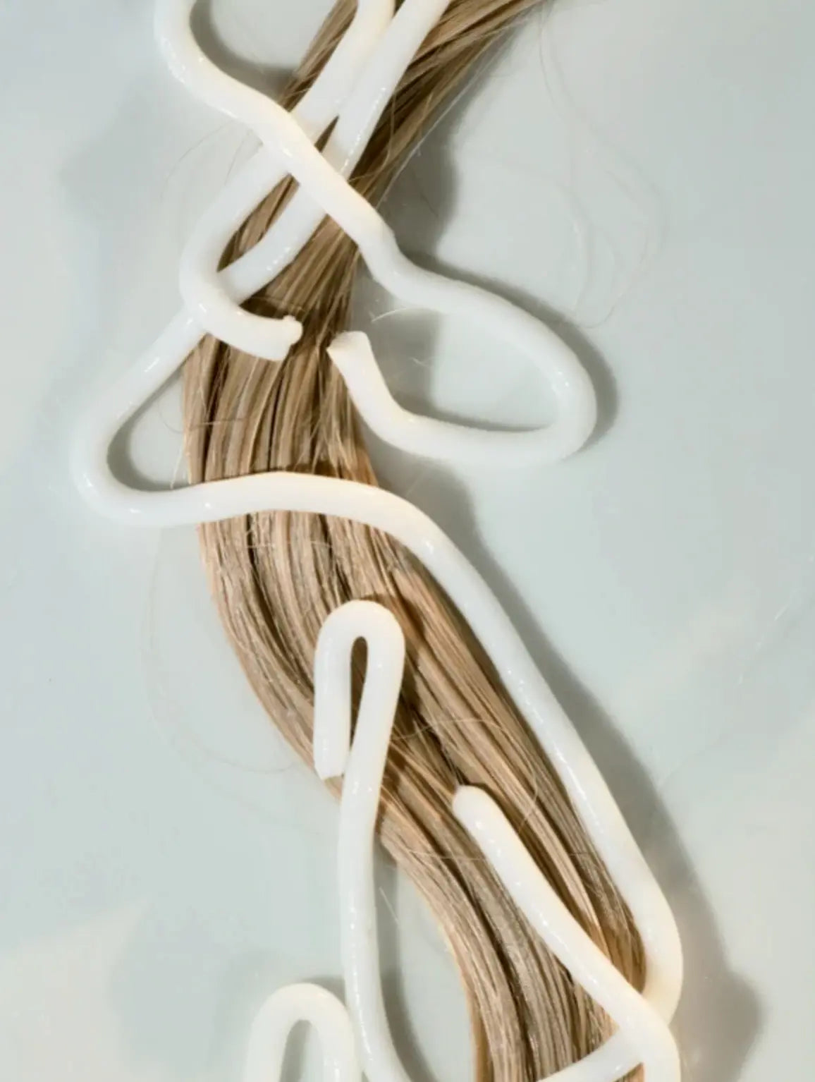 A close-up of a bundle of light brown hair extensions or a wig section wrapped with a thick, glossy white adhesive, set against a clean, pale gray background.