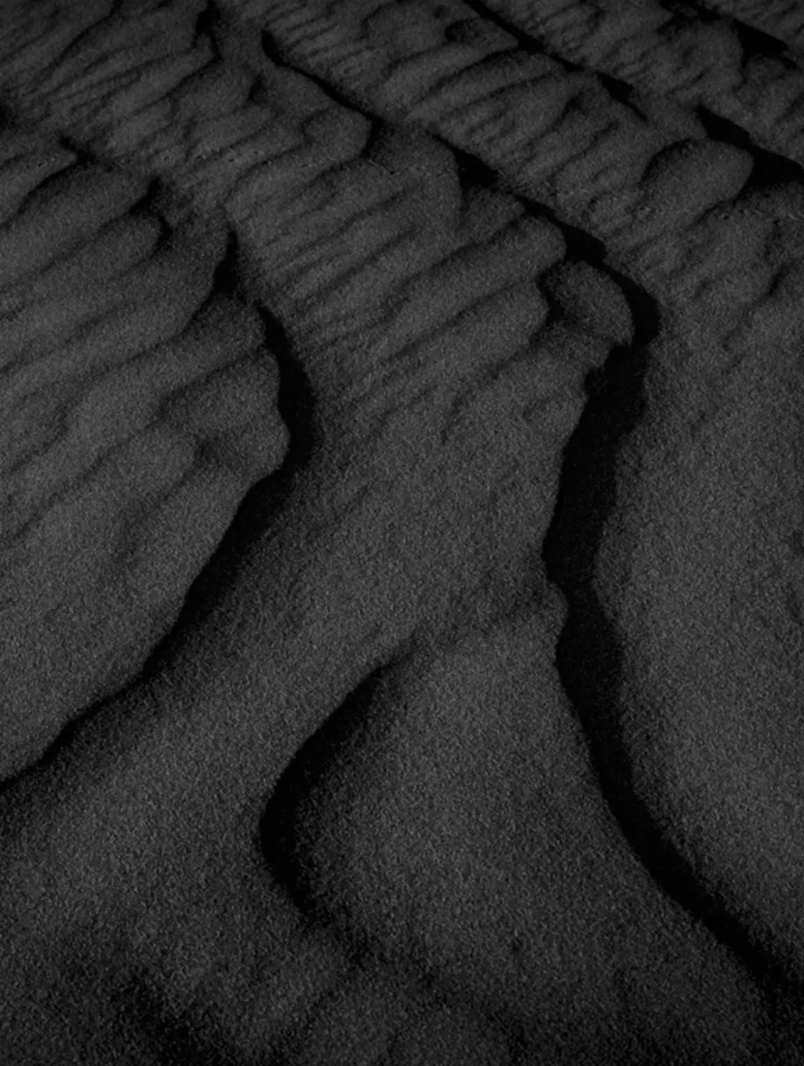 A close-up of dark textured material with flowing, ribbed ridges and deep shadows, resembling black fabric, leather, or a similar material swatch.
