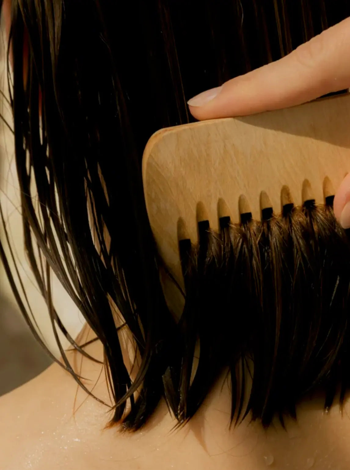 A close-up of wet dark hair being brushed with a light natural wood hairbrush by a person's hand.