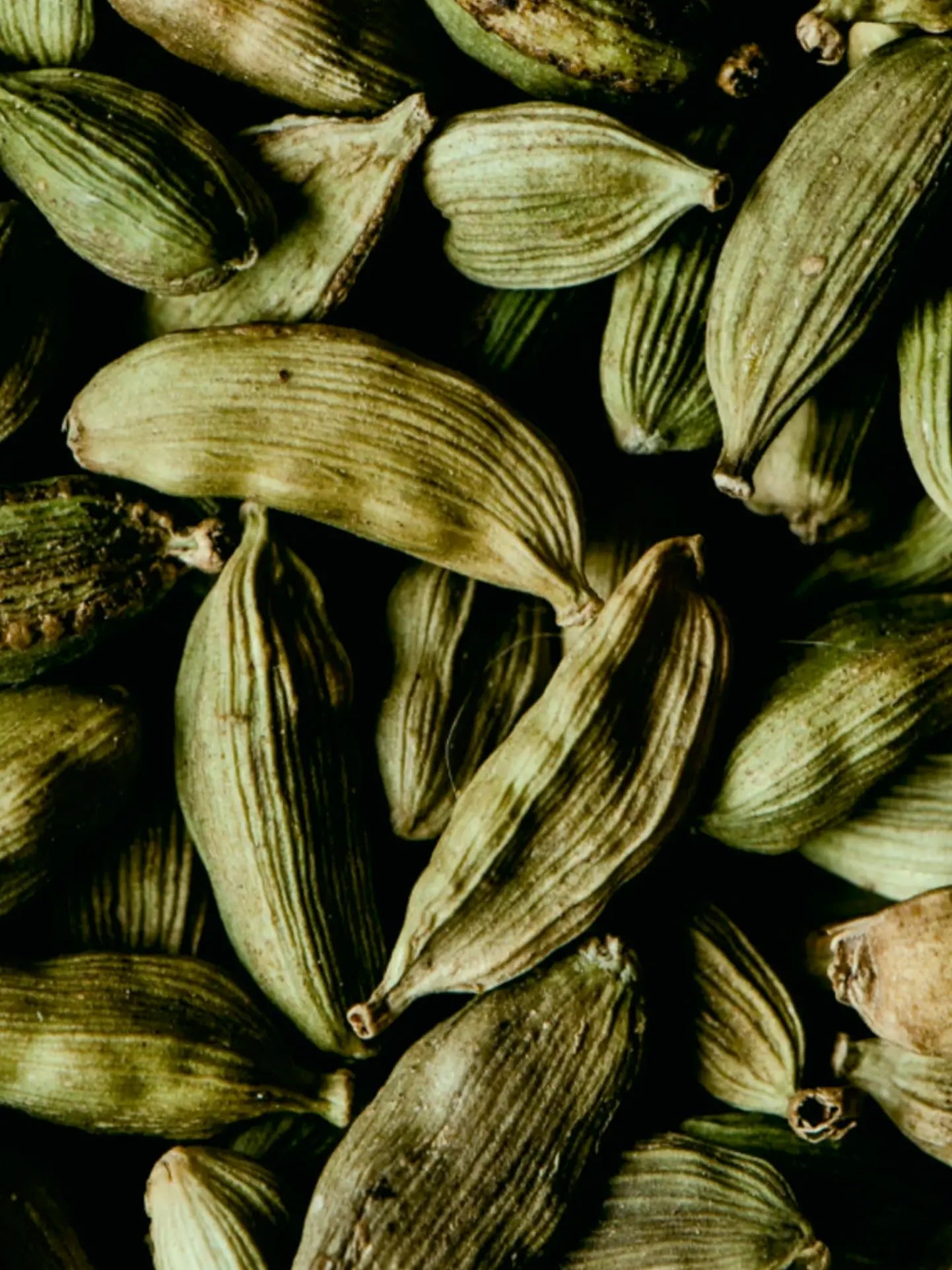 A close-up pile of dried green cardamom seeds with visible ribbed texture against a dark background.