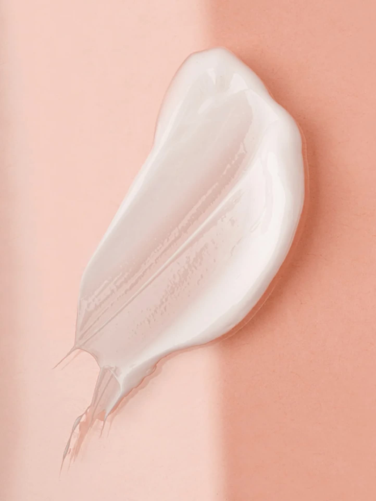 A thick, creamy white cosmetic cream swatch applied in an irregular smear on a smooth, solid peach-pink background.