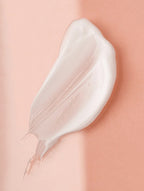 A thick, creamy white cosmetic cream swatch applied in an irregular smear on a smooth, solid peach-pink background.