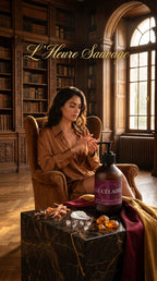 A woman sits in a brown armchair in a warm, book-filled library setting, applying a product from a pump bottle placed on a dark marble table. The bottle label reads 'LE CÉLAIRE' and the image has the text 'L'Heure Sauvage' at the top.