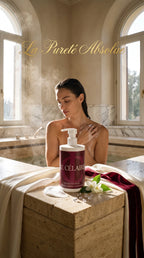 A woman sits in a steamy stone bathroom with arched windows, applying cream to her shoulder. In the foreground, a bottle of La Pureté Absolue Célaines intensive skin body wash rests on a stone ledge with a towel and white flower prop.