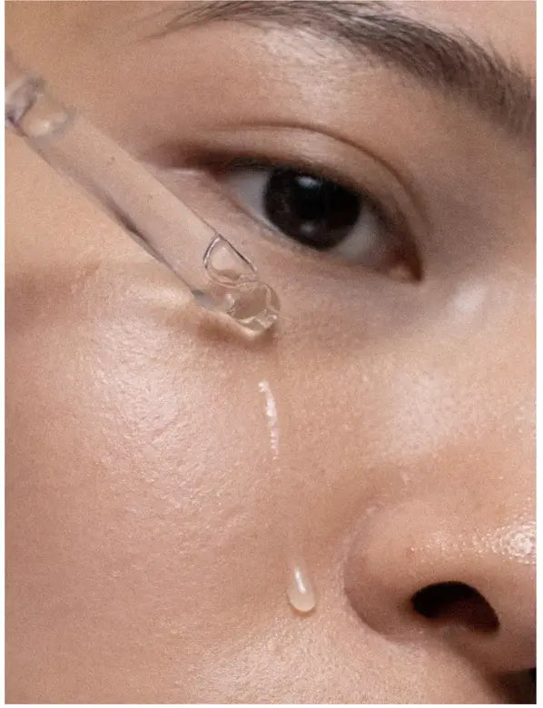 Close-up of a person's eye area with a clear glass dropper applying a transparent liquid serum, with a drop falling onto the skin.
