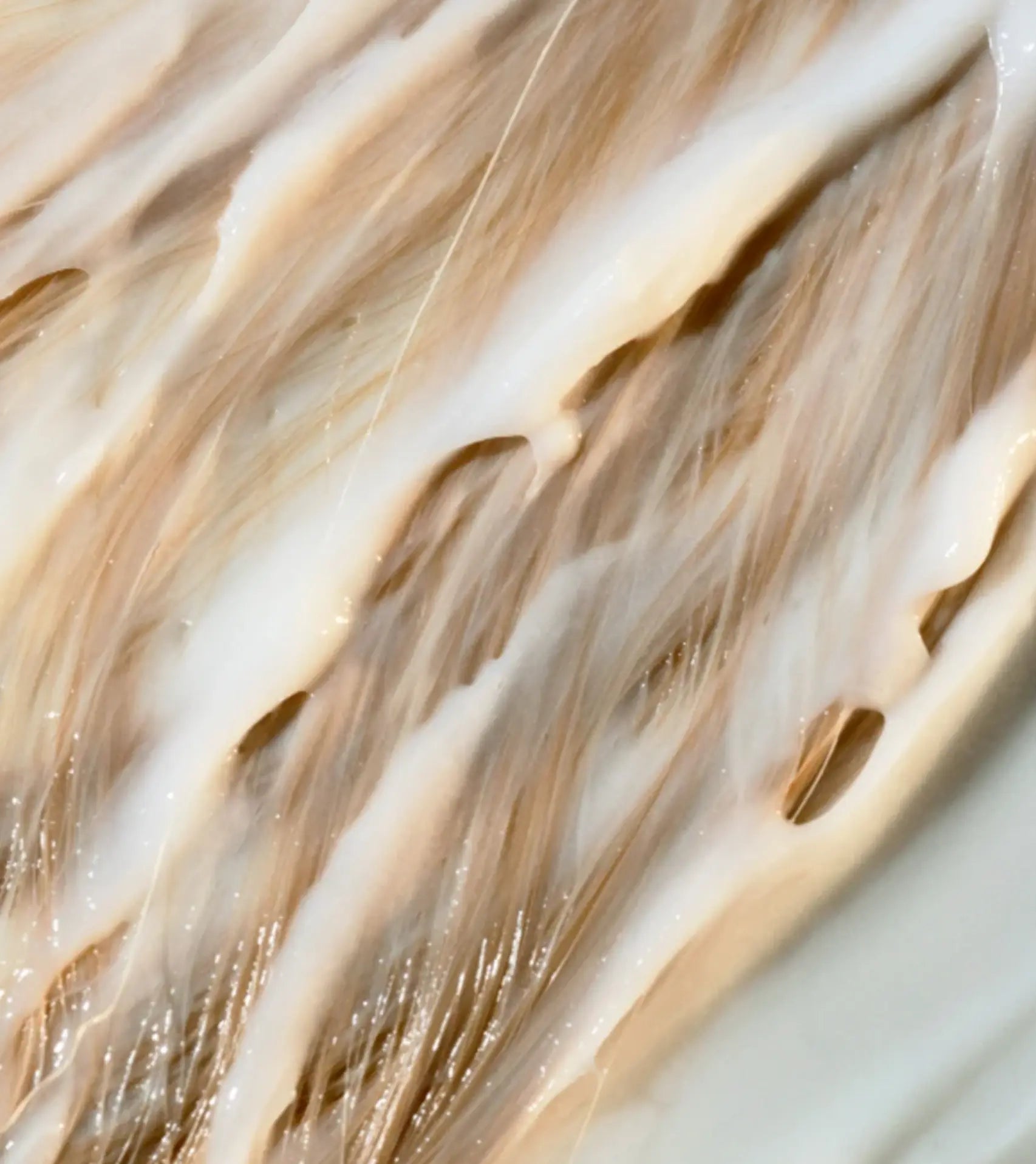 Close-up of glossy, wet hair strands coated with a creamy white styling product, showing warm beige and light brown tones.