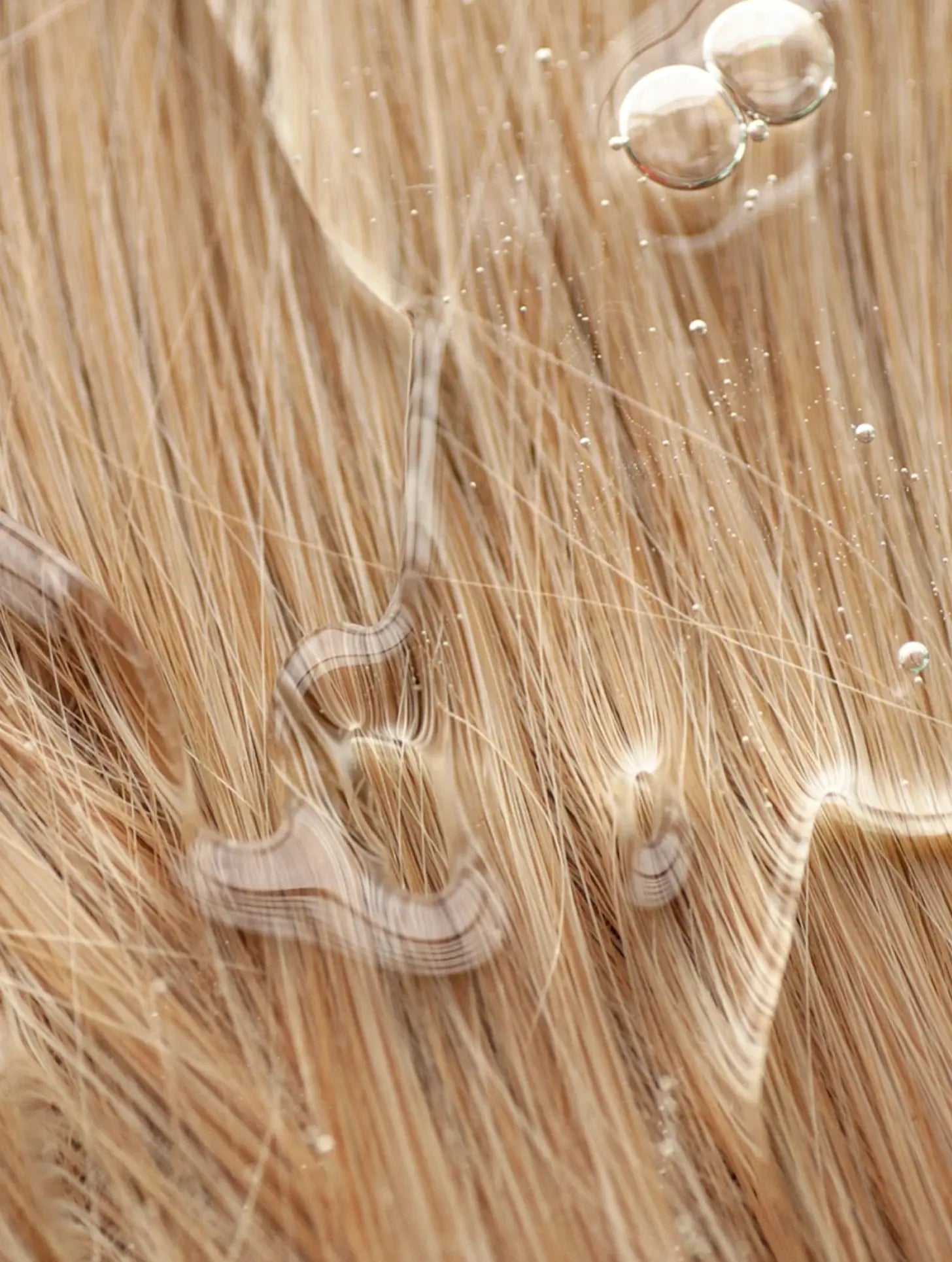 Close-up of light blonde hair with water droplets and oil sheen, showing strands and glossy reflections.