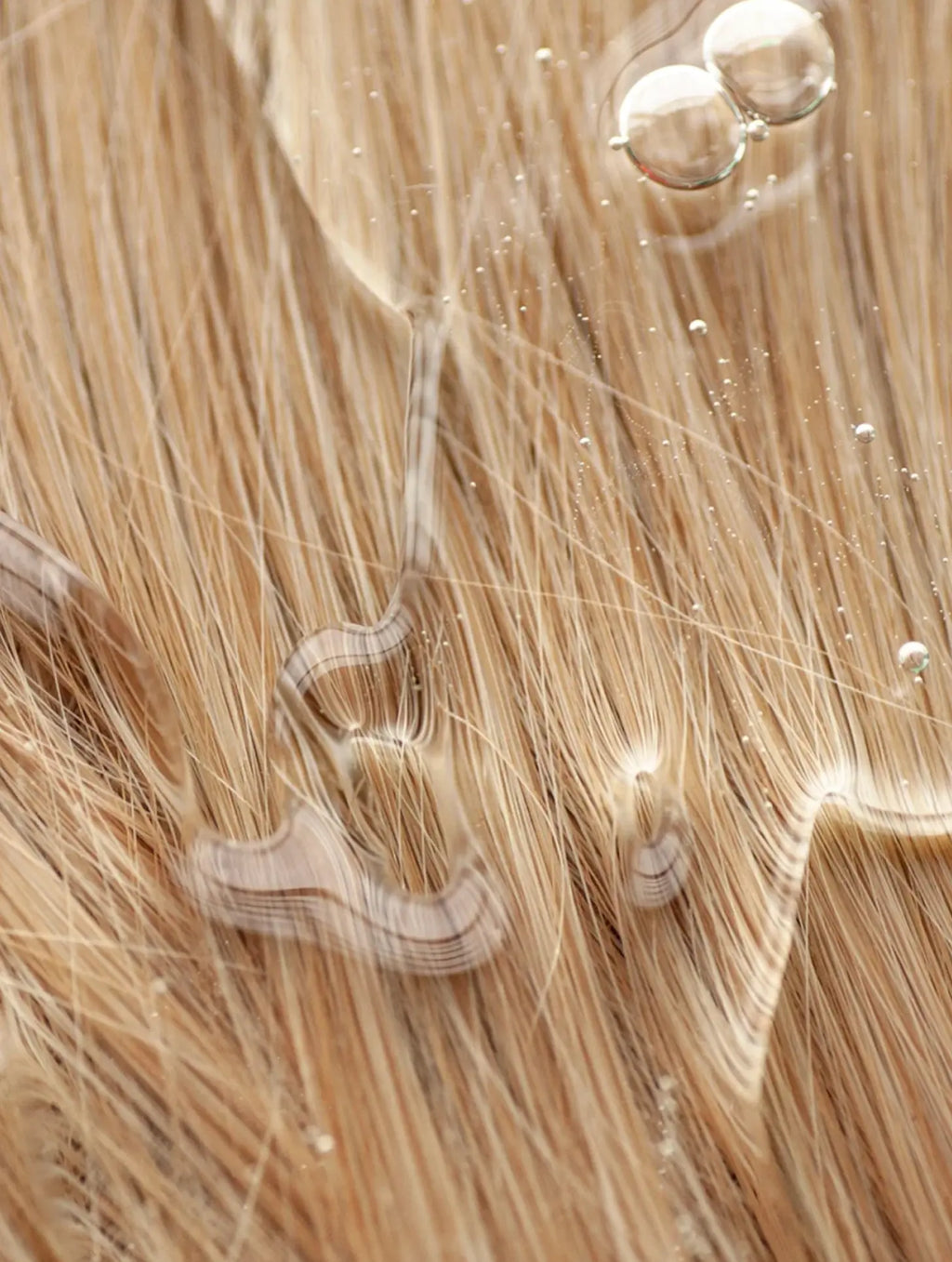 Close-up of light blonde hair with water droplets and oil sheen, showing strands and glossy reflections.