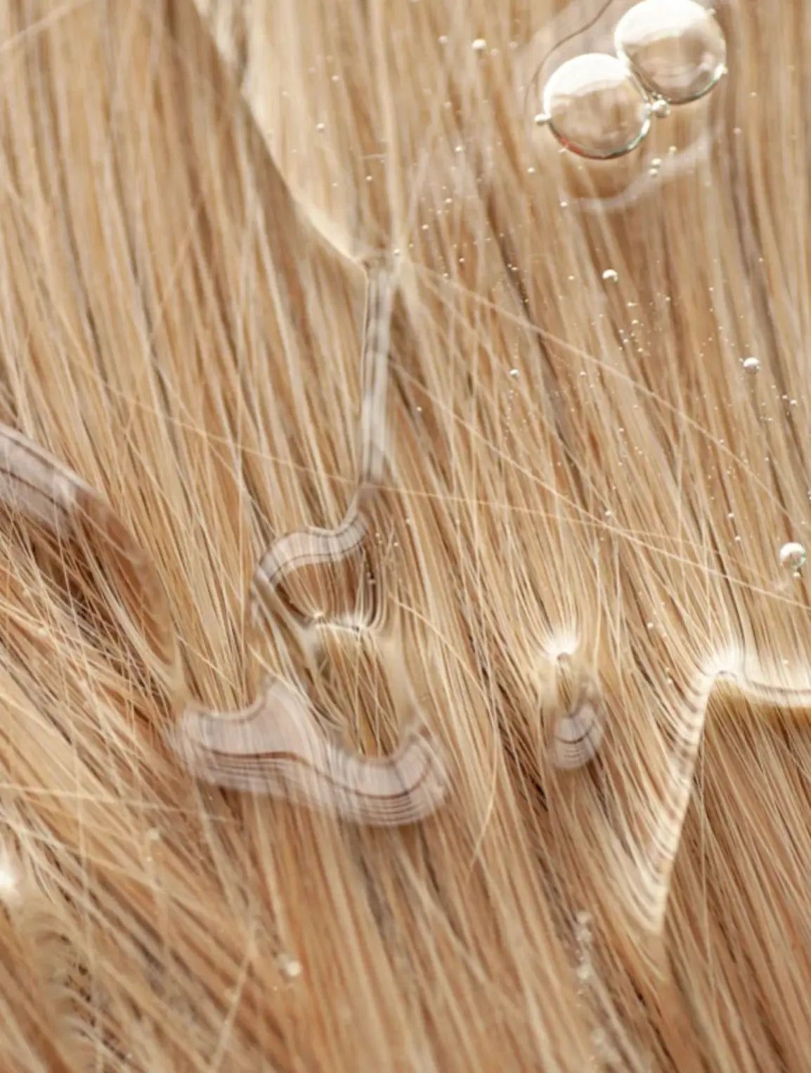 Close-up of light golden-blonde hair with water droplets and a glossy sheen, showing texture and shine in macro detail.