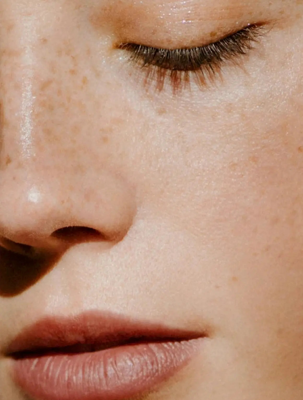 Extreme close-up of a person's face showing natural skin texture, freckles, closed eyelashes, and lips under warm directional sunlight.