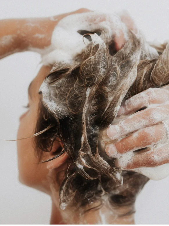 Woman using Sensitive Scalp Shampoo for all Hair Types, applying gentle foamy cleanser to soothe scalp.