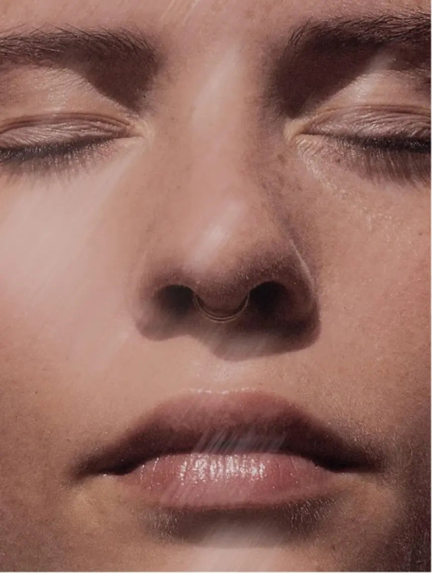 Extreme close-up of a person's face with closed eyes, showing skin texture, pores, and a small metallic ring on the nose.