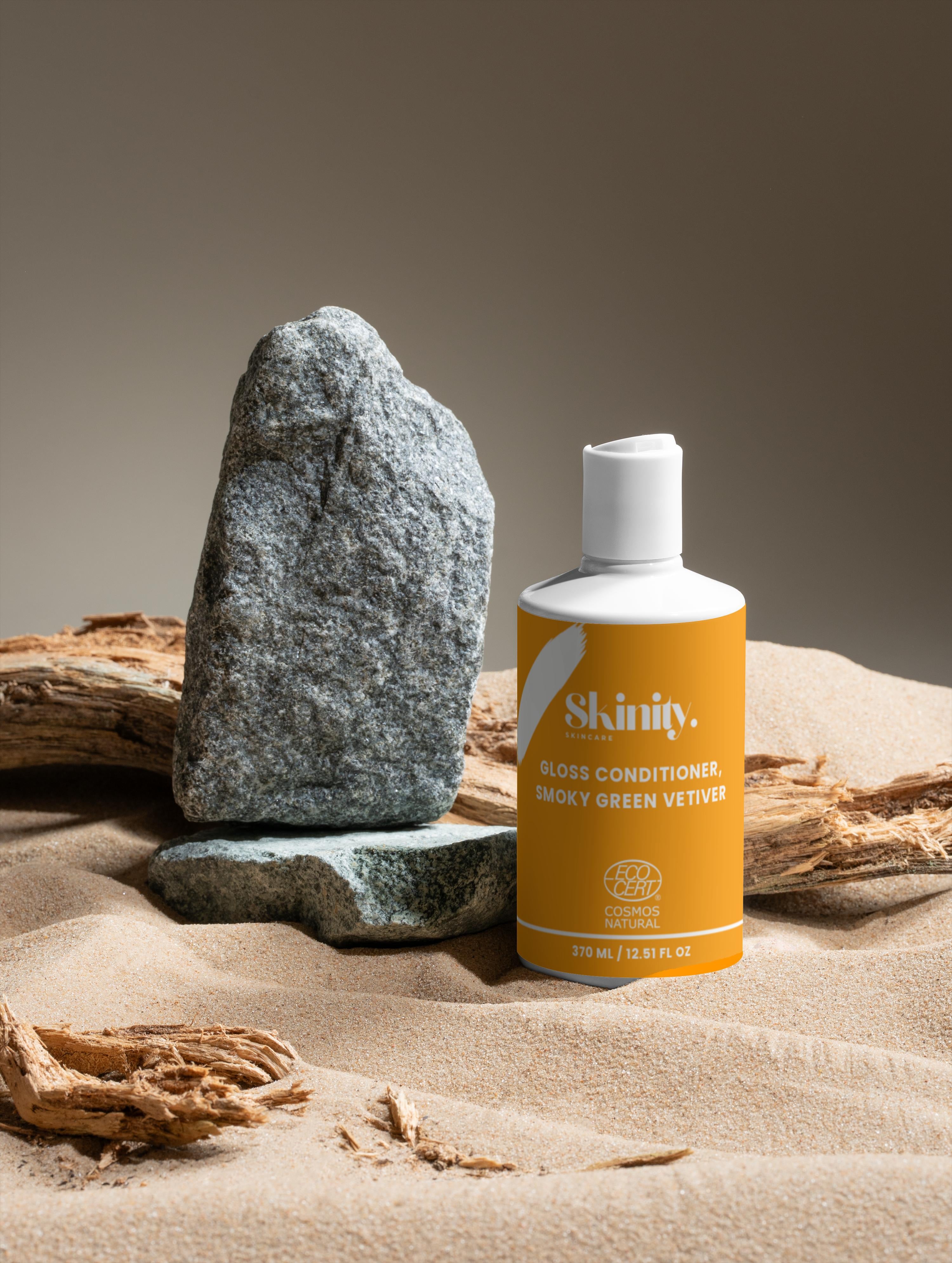 Gloss Conditioner Smoky Green Vetiver bottle on sand with stone and driftwood, promoting shiny, smooth hair.